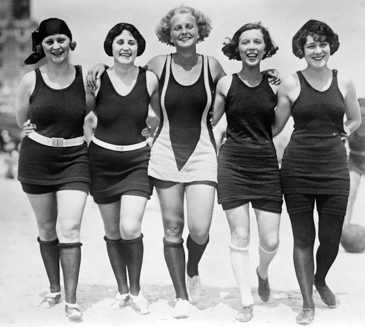 Five women walking arm-in-arm on the beach wearing wool bathing suits, 1925