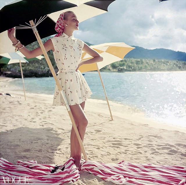 Model standing under beach umbrella wearing summer dress look; pleated short shorts and matching sleeveless shirt, both in a light floral pattern. 1954