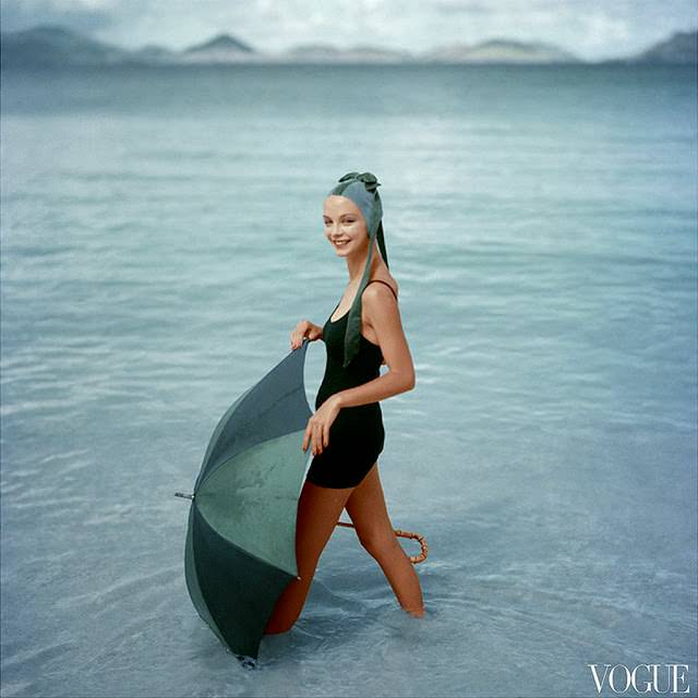 Gretchen Harris, standing in ocean with open green and blue umbrella, wearing knitted black maiilot of DuPont and water-velvet cap , 1957