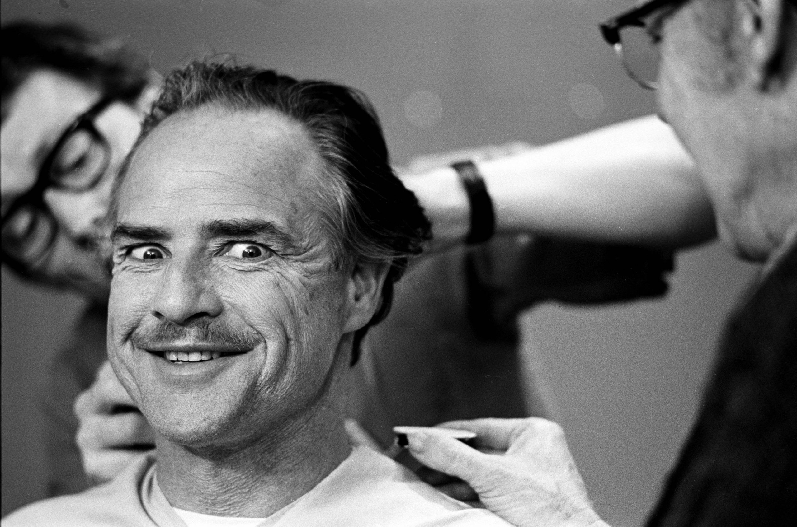 Marlon Brando grinning devilishly while getting his make-up done during the filming of The Godfather in New York, 1971