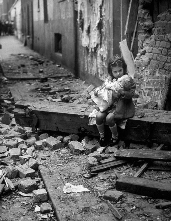 A little girl holds her doll in the rubble of her bomb-damaged home. England. 1940.