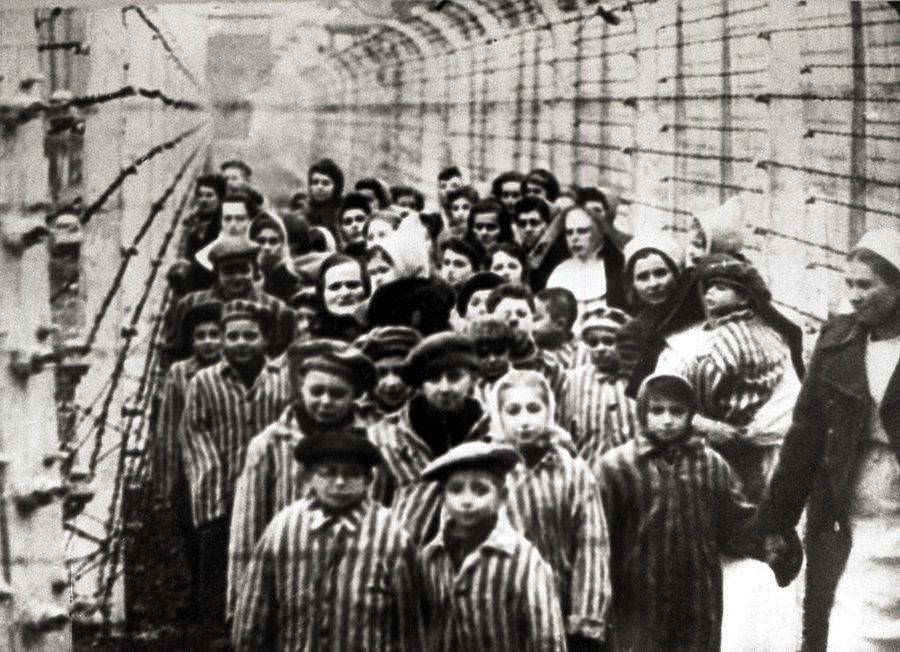 Jewish children, survivors of Auschwitz, stand with a nurse behind a barbed wire fence. Poland, 1945.