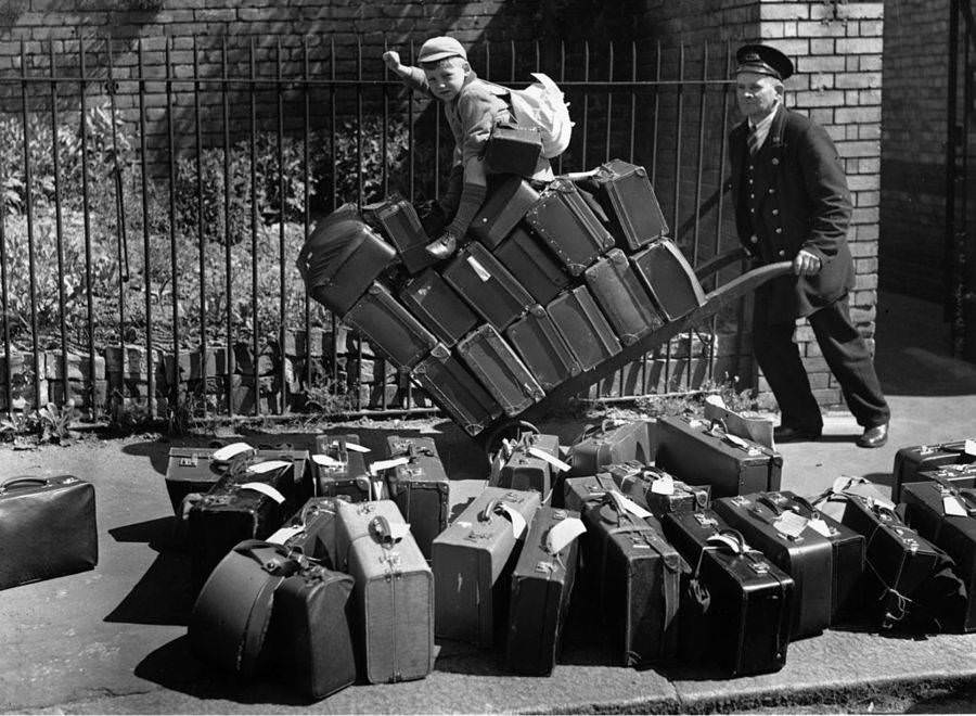 A porter pushes the luggage of evacuees bound for Wales on a trolley at a London railway station, with a young boy perched on top of the suitcases, 1940.