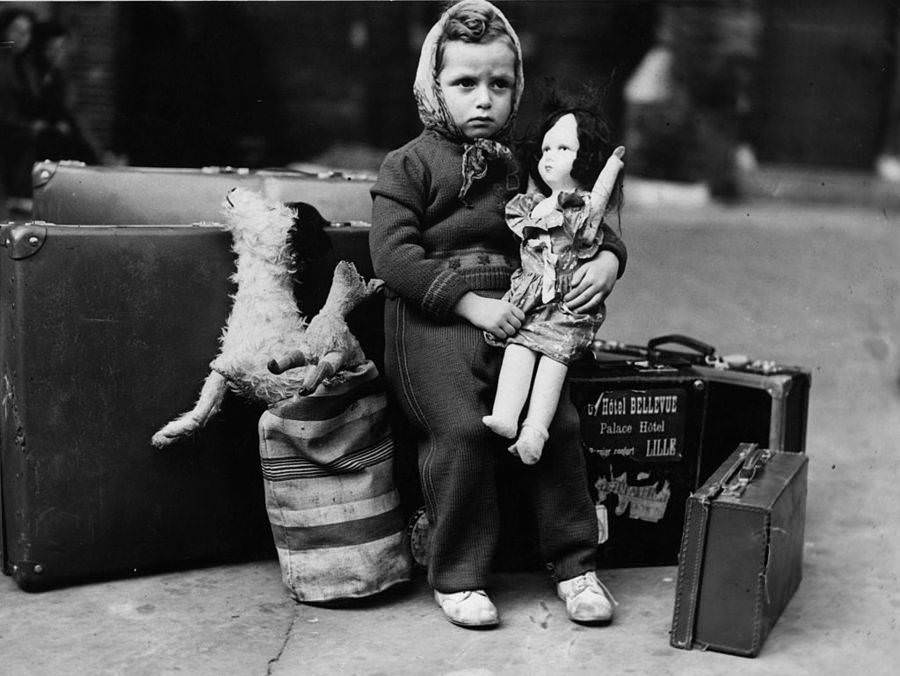 A little girl waits nervously with her doll and luggage before leaving London for her billet, 1940.