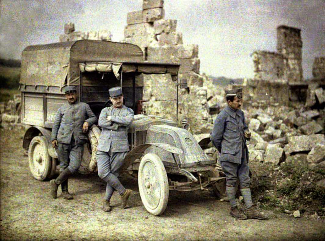 Three French soldiers take a break in front of a heavily damaged building with their small truck, 1917.