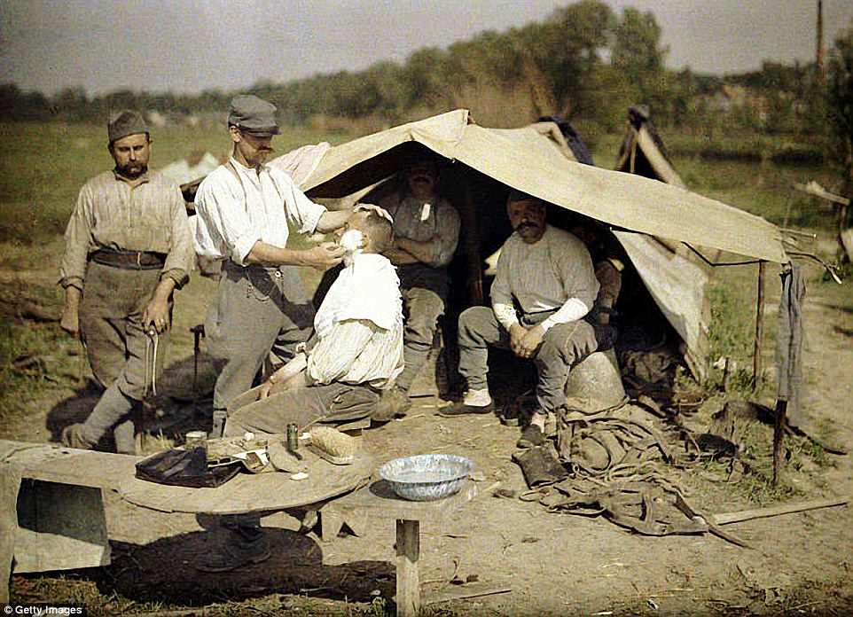 A soldier is shaved by a barber in a French military encampment in Soissons, while two soldiers wait under a tent, in 1917
