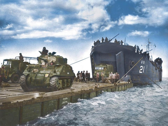 The USS LST-21, manned by the U.S. coastguard unloads British Army tanks and trucks onto a Rhino barge in the opening hours of their invasion of Gold Beach.