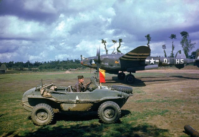 A P-38 fighter plane sits in the background as the pilot arrives in a captured German vehicle, France