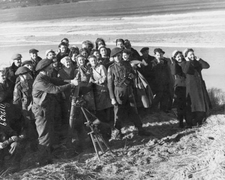 A group of mostly women war workers stand, many with their fingers in their ears, on the sand of a coastal test range to watch as several soldiers launch mortar bombs, 1943.