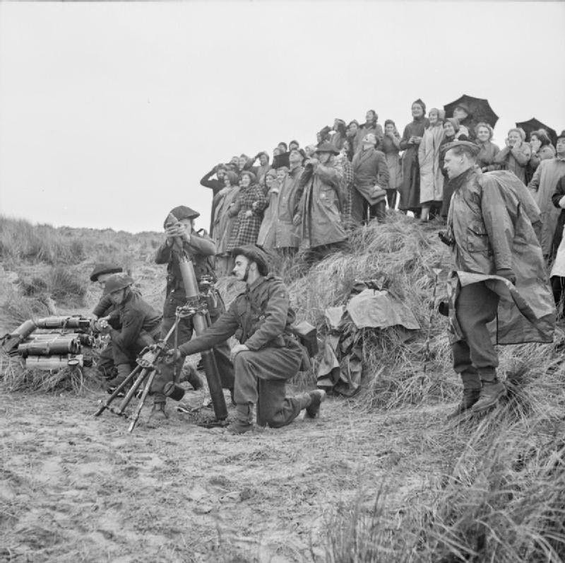 A group of war workers stand, on a grassy mound at a coastal test range to watch as soldiers launch mortar bombs from the beach immediately below them, 1943.