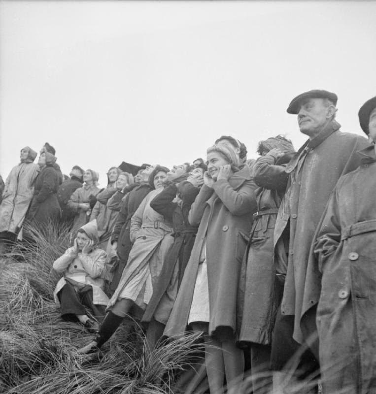 A group of war workers stand in a row on a grassy hill on a coastal test range to watch mortar bombs being launched, 1943.