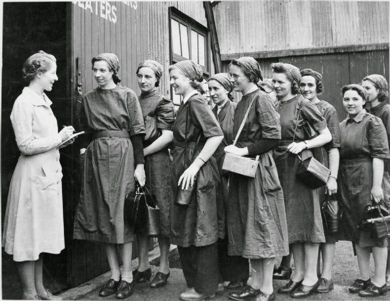 A group of new recruits dressed in overalls and head scarves and carrying their gas masks are met by the female supervisor at Slough Training Centre as they enter the workshop for the first time, 1941.