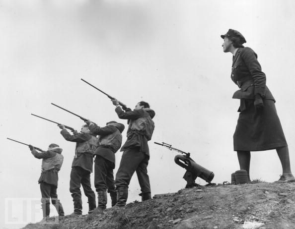 Pilots of a Spitfire fighter squadron, wearing their life-vests to be ready to take off at any time, take part in a clay pigeon shoot. An officer of the Women's Auxiliary Air Force assists them by operating the clay pigeon trap.