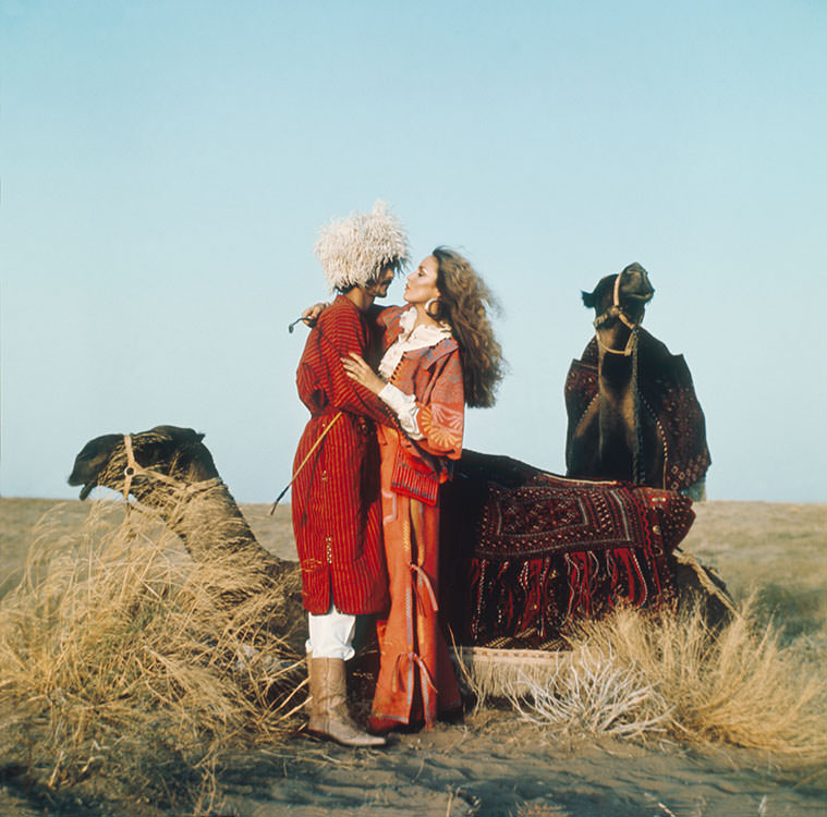 Jerry Hall embracing a man surrounded by two camels, 1975