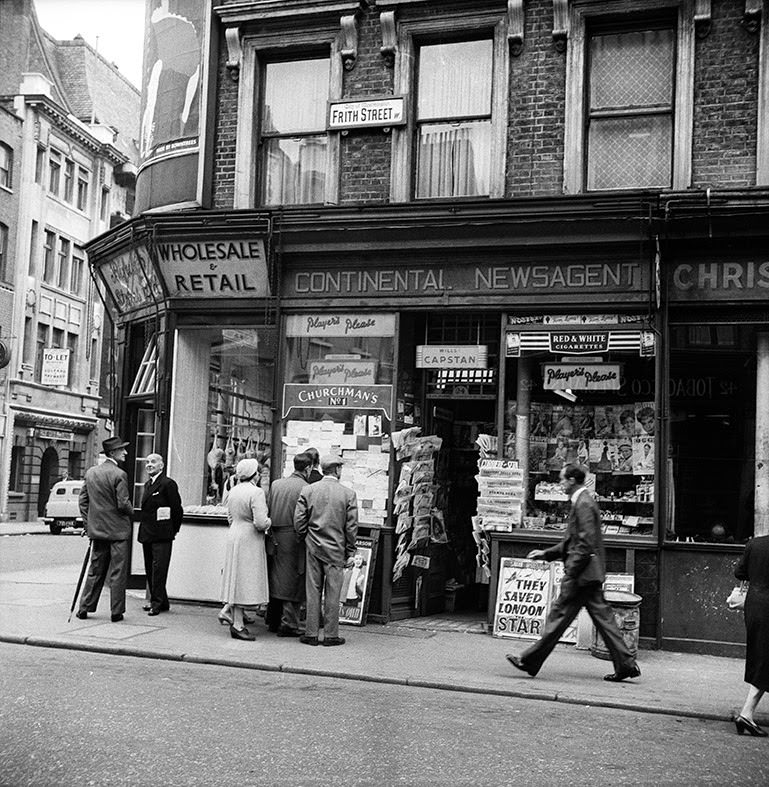 People look at notices outside a newsagent's shop on Frith Street, where a newspaper board reads: 'They saved London', 1956