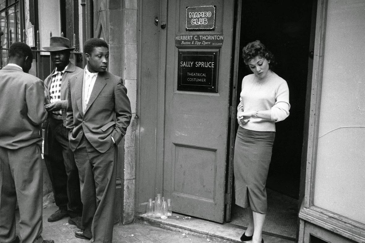 Andria Loran, a model who was democratically elected Queen of Soho 1956, stands in the doorway of the Mambo Club, keenly noticed by an onlooker