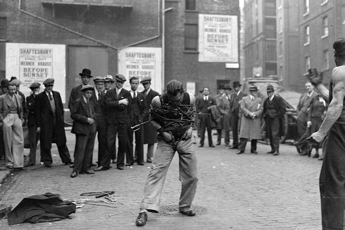 A small crowd gathers to watch a man trying to extract himself from chains on a street in Soho, 1937