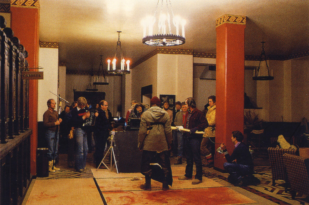 Filming Dick Hallorann’s death scene on the lobby set. Stanley Kubrick composes the insert shot of the axe striking Hallorann’s chest. A special effects artist kneeling to the right of Jack Nicholson holds a bag of fake stage blood, with tubing to deliver the blood running up the handle of the axe.