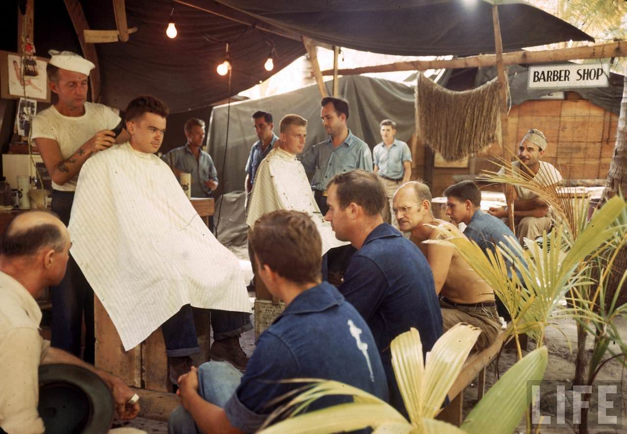American servicemen waiting to get haircuts at a makeshift island barber shop during WWII.