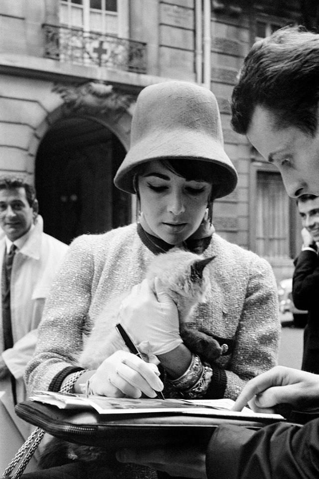 While returning to Paris from a trip to Russia, Taylor proved herself capable of simultaneously signing an autograph and stroking the nape of a mysterious cat, 1961