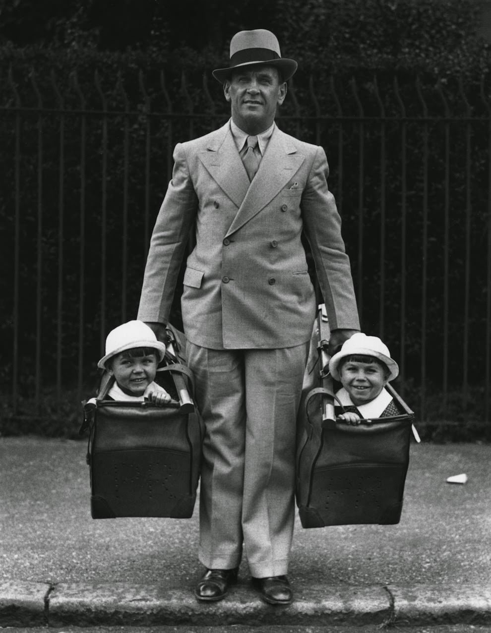 A man carrying twins, two 36-year-olds who are part of a circus act, in his bags, 1935
