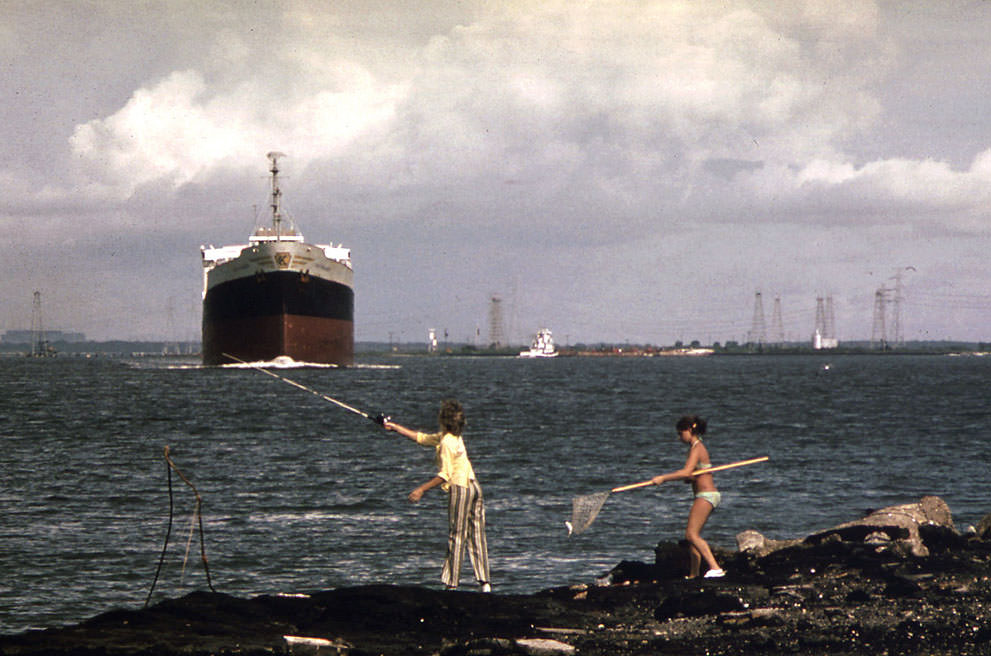 A freighter moves slowly up the Houston ship channel as girls fish from the shore, September 1973. Oil derricks are visible in the background. (Blair Pittman/NARA)
