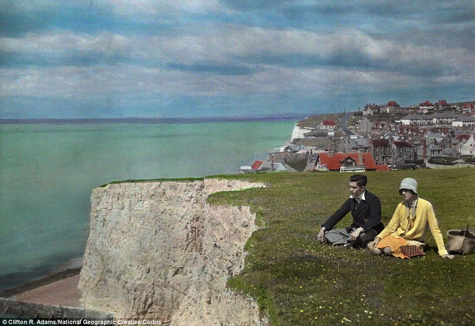 Two young adults sit in the grass by a cliff near water and the village of Rottingdean in East Sussex in 1931