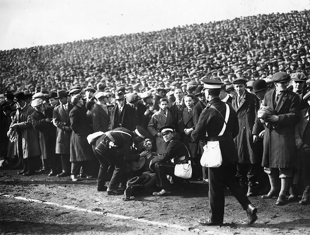 A woman who has fainted receives medical attention on the pitch sidelines during the FA Cup Semi-Final between Sheffield Wednesday and Huddersfield Town at Old Trafford, 22nd March 1930
