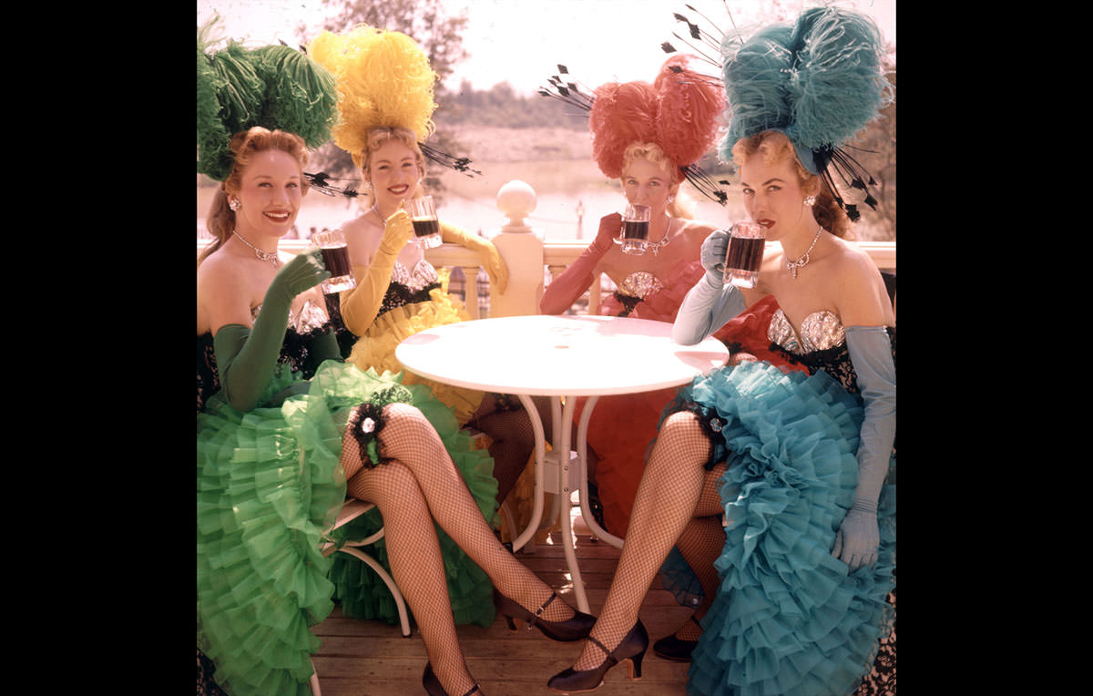 Showgirls in costume sit at an outdoor table and drink from mugs at Disneyland in July 1955