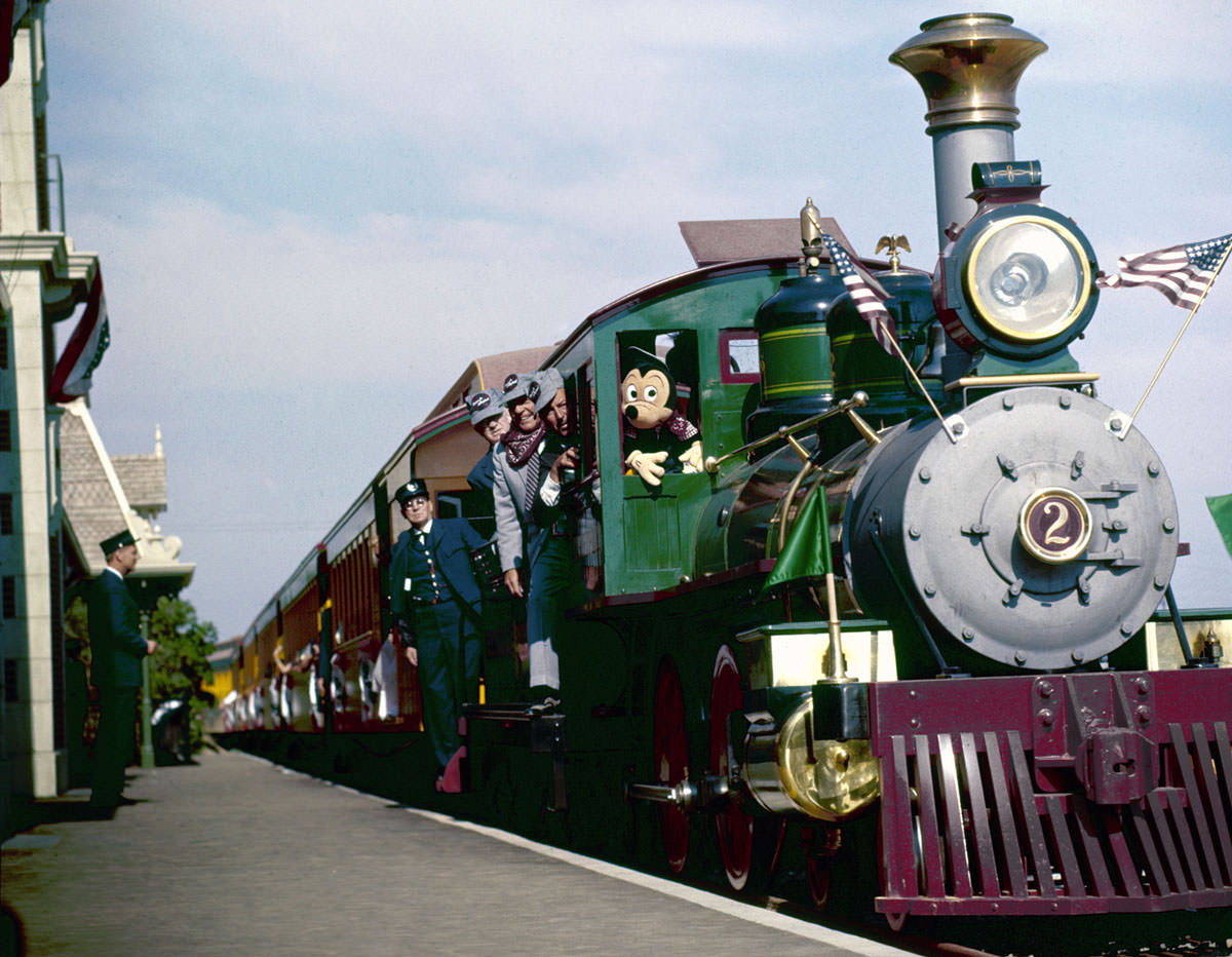 Mickey Mouse rides in the locomotive cab of a train (with Walt Disney just to his left) as it arrives at a station in Disneyland on July 18, 1955