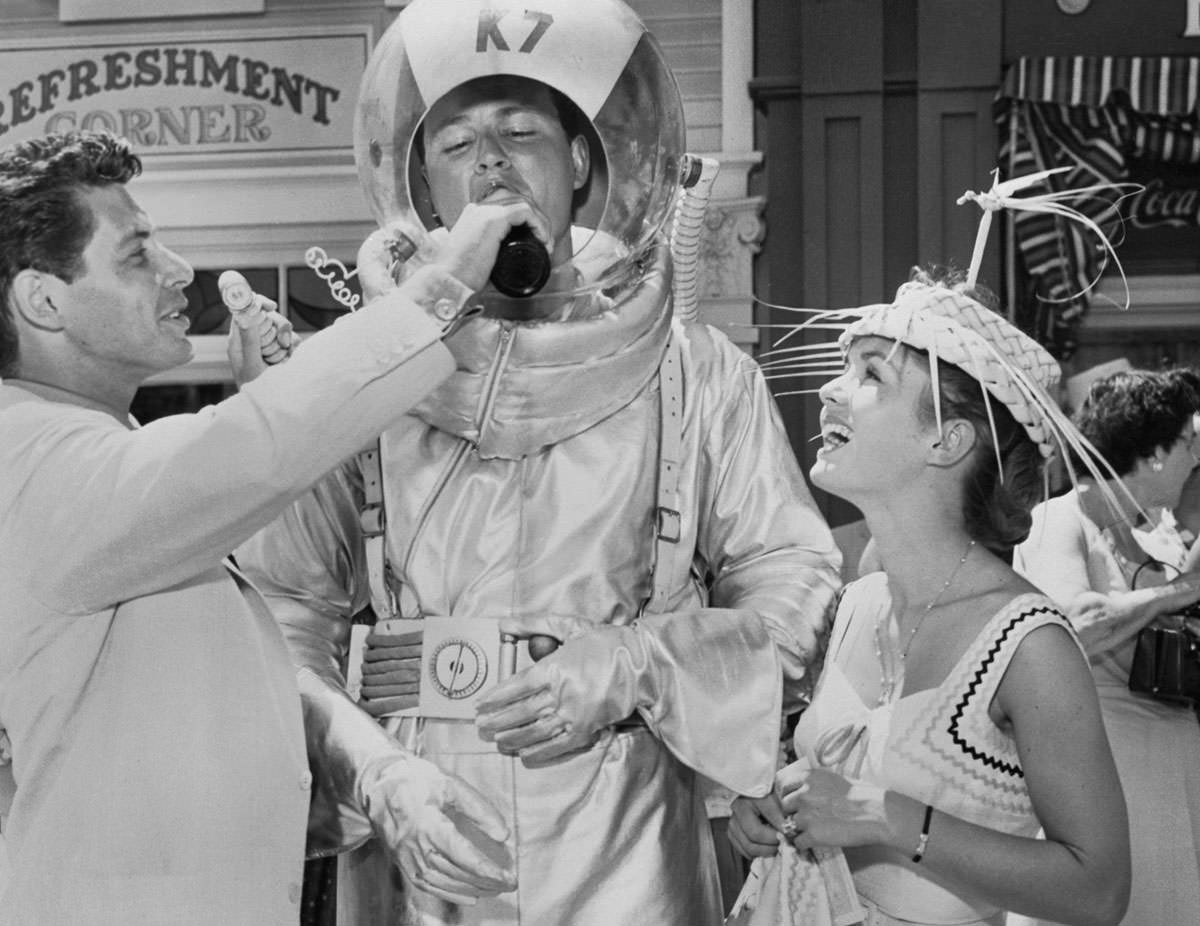 Original caption: "Eddie Fisher (left) was a host at the opening of Walt Disney's 'Disneyland,' where he served on the Coca-Cola refreshment corner. Shown are Eddie Fisher as he gives a drink to aluminum-clad spaceman Don MacDonald, as Debbie Reynolds looks on