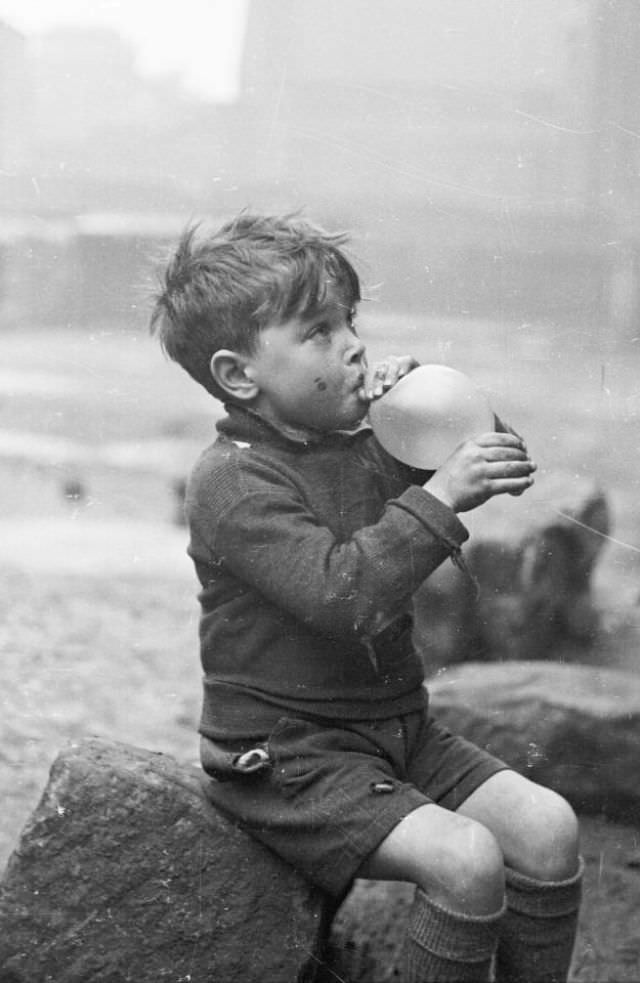 A young boy sits on a stone and attempts to blow up his balloon in the Gorbals