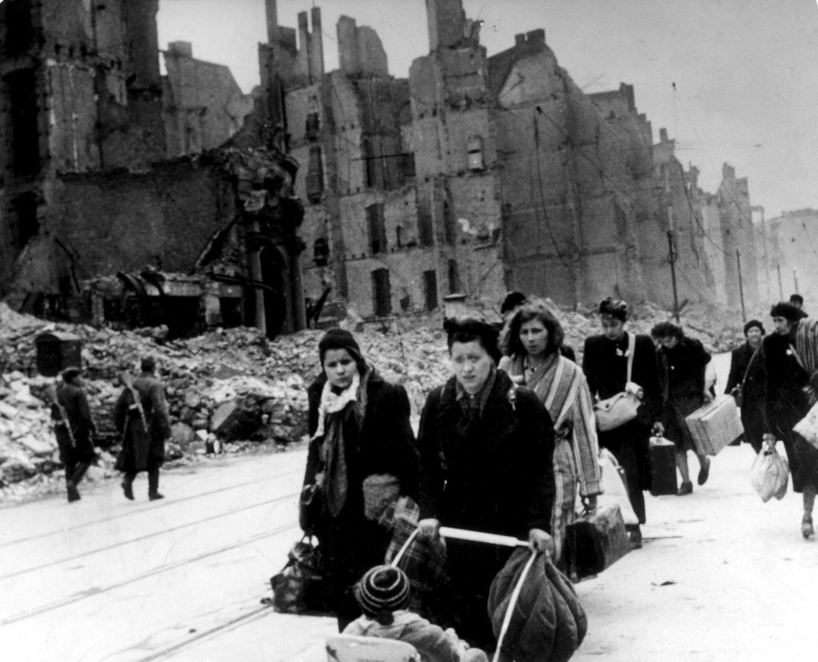 A stream of refugees and people who have been bombed out of their homes moving through destroyed streets in Germany in 1945, after end of war. On the left, two Soviet soldiers can be seen patrolling.