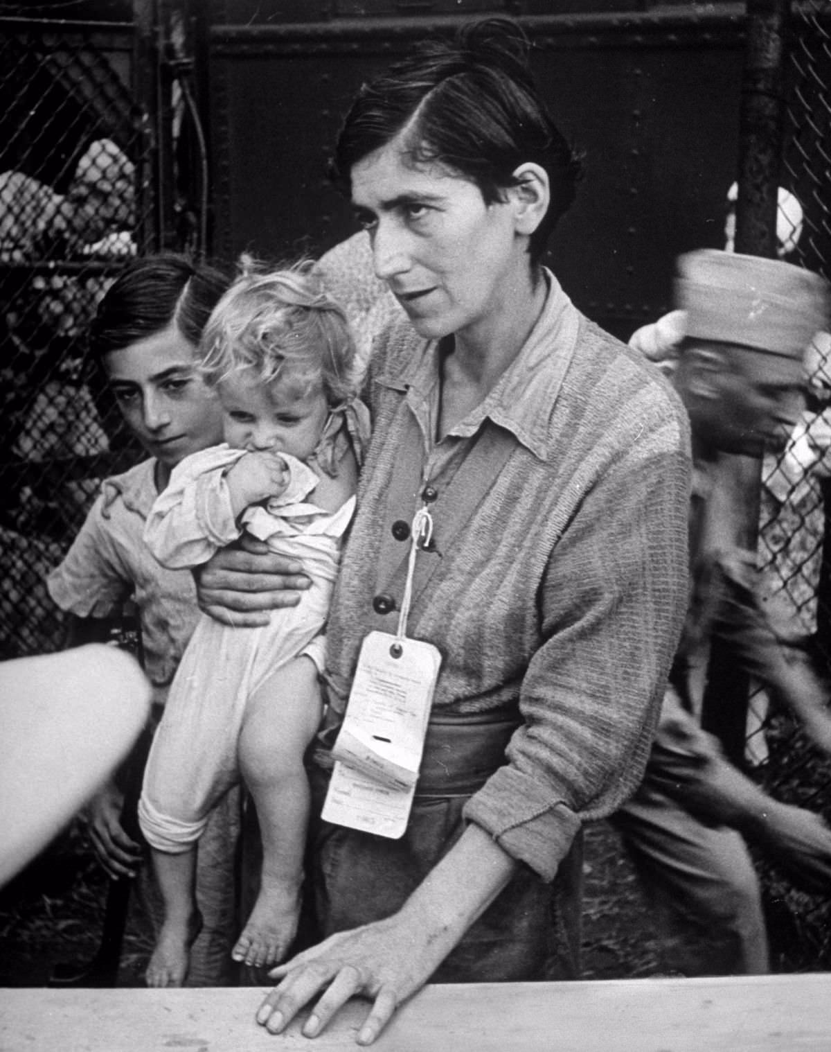 With her baby in her arms and her young son tagging behind, Mrs. Eva Bass comes through gate to her new home at Fort Ontario.