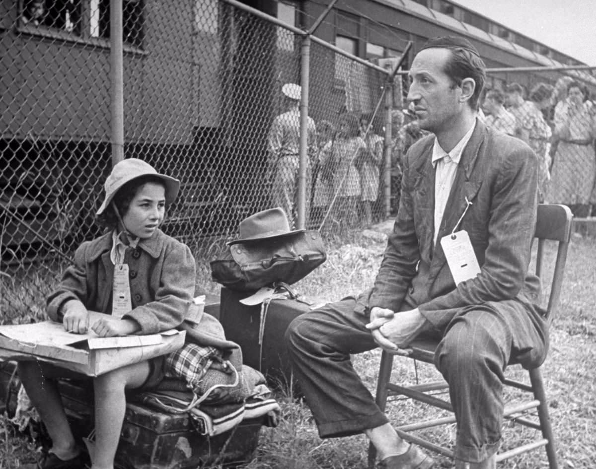 At camp gate Victor Franco from Tripoli waits with his daughter for his wife who is pregnant and rode in hospital car. He was afraid he might lose track of her, even though attendants told him he could see her in the hospital. He said he would wait right where he was.