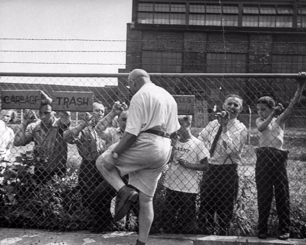 Americans from Oswego line high wire fences of Fort Ontario to stare curiously at the refugees. Here one of the foreigners walks over to talk with them. He scratches his bare leg while explaining that he has a son in the U.S. Army.