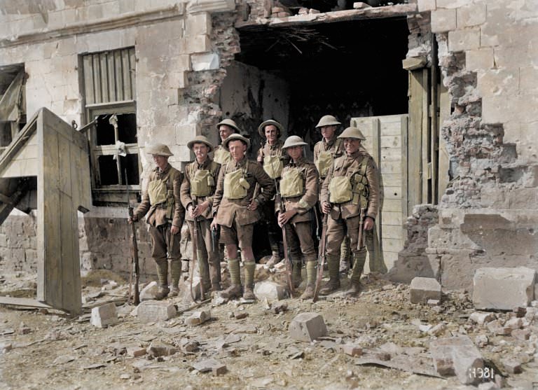 A group of eight British soldiers stand next to a blown-out building. They smile as they pose for the camera wearing their helmets.