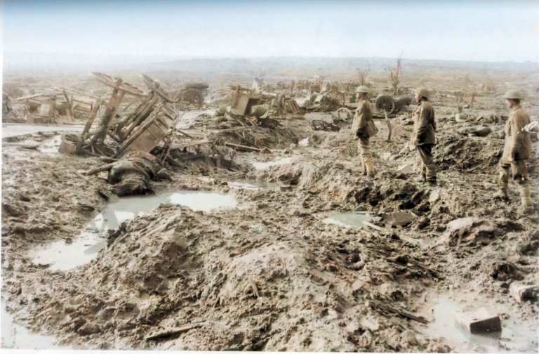 Three soldiers look out across a battlefield where wagons are upturned and destroyed and craters break up the mud.