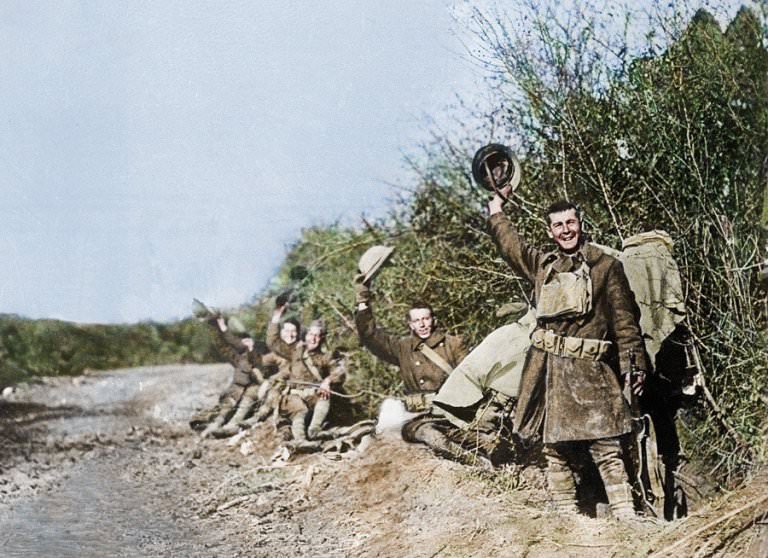 A group of troops wave their hats as they pose for a camera on the edge of a road next to some hedgerows on Armistice Day.