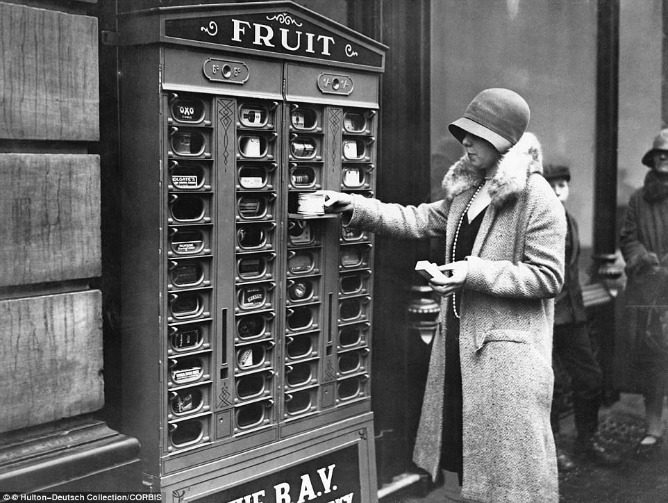 A woman in London is able to continue her grocery shop thanks to a vending machine which says it dispenses fruit but seems to offer kitchen cupboard essentials such as Oxo cubes, tins of food, matches and Colgate products, 1920.