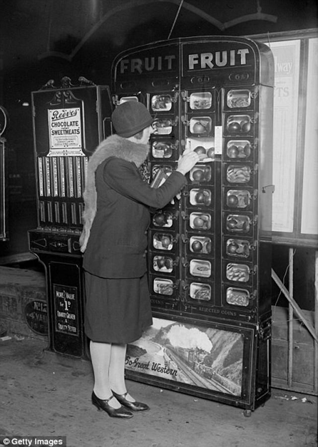 A woman buys fruit from a coin operated machine at Paddington Station in London, ca. 1920s.