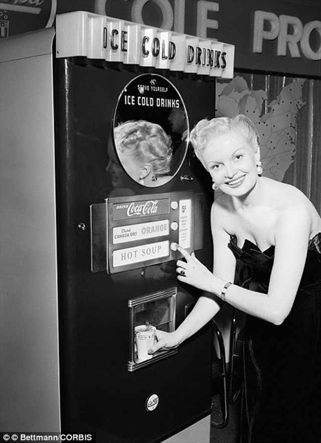 A woman demonstrates a multi functional vending machine that dispenses both cold drinks including Coca Cola, Canada Dry and orange juice along with hot soup, ca. 1950s.