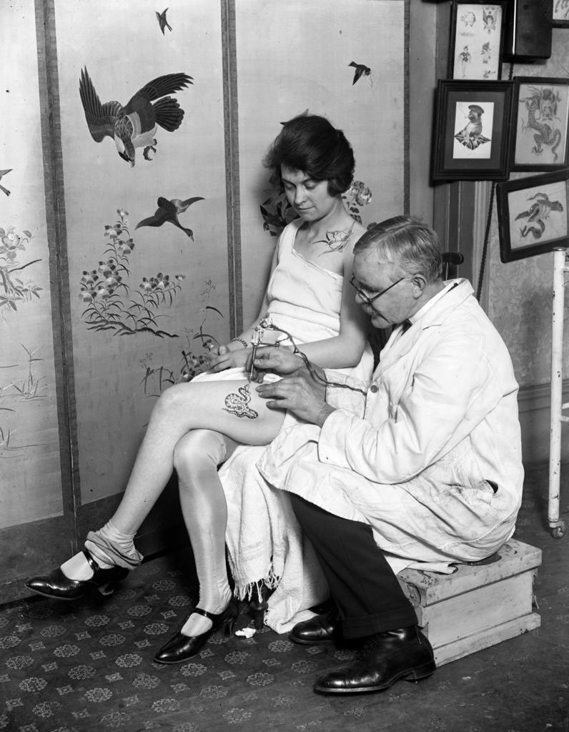 A woman having an image of a snake tattooed onto her thigh by George Burchett, 1928.