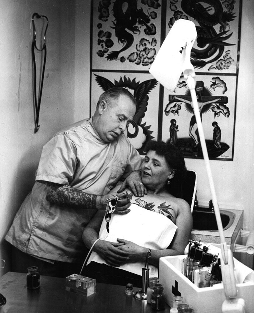 A tattooist painting a bluebird on a woman’s breast, 1965.