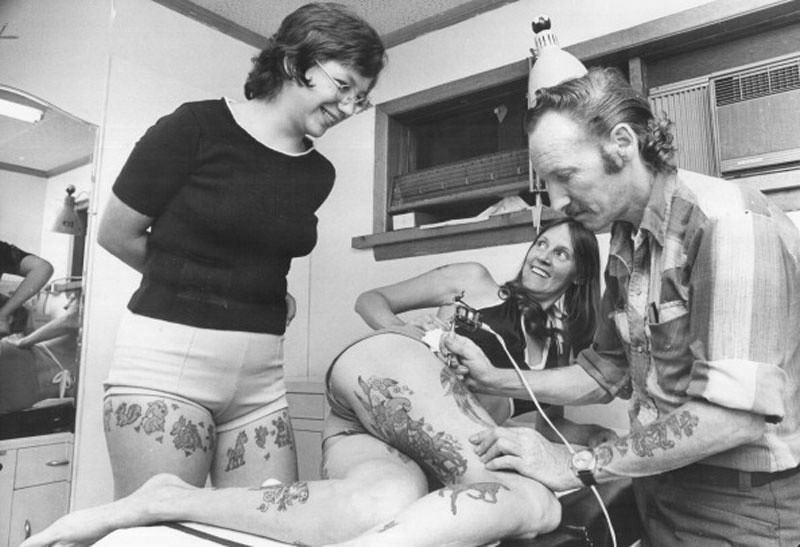 Beachcomber Bill’s wife; Brenda; smiles from table as he shows how he applies tattoo, 1975.