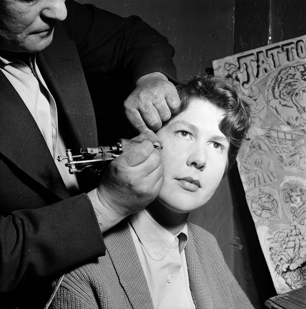 A tattoo artist paints a permanent beauty spot on the cheek of a woman at his workshop in Copenhagen in 1956.