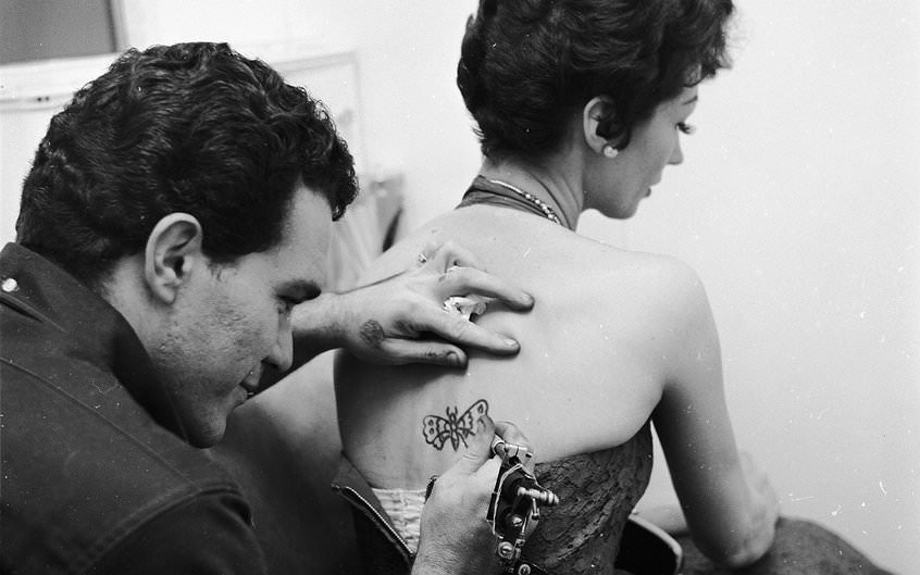A tattoo artist tattooing a butterfly onto Lulubelle Hostein's back, ca. 1955