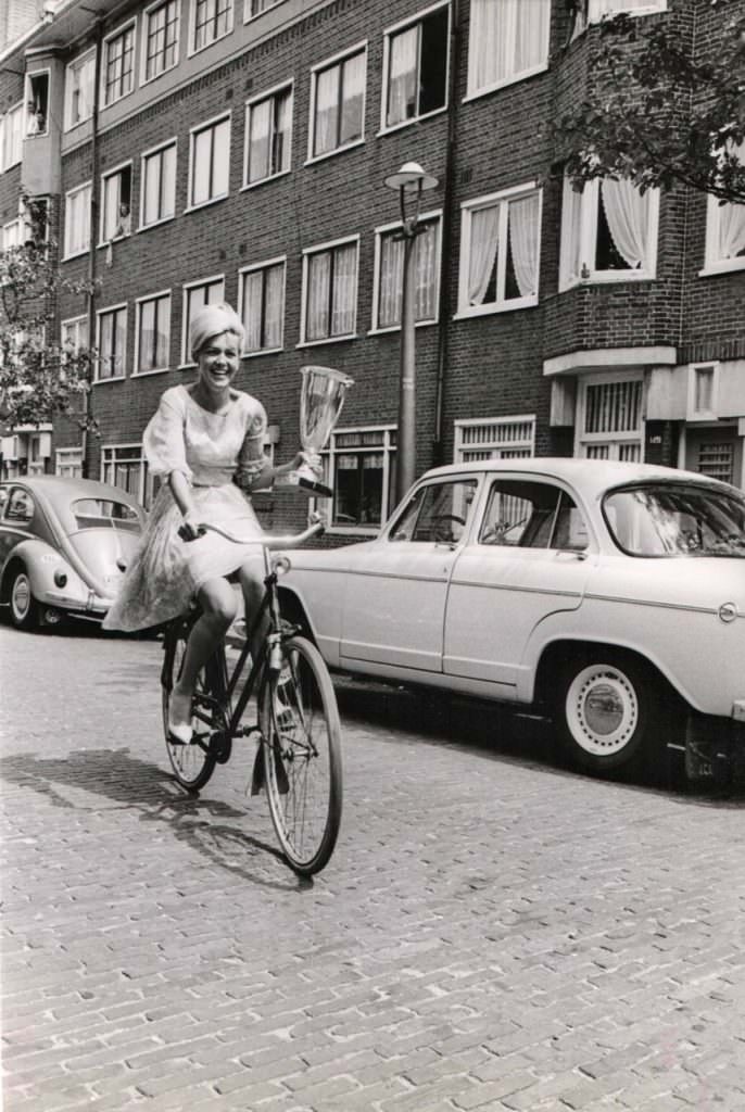 Elly Koot riding a bike, wields the 1964 Miss Europe trophy.