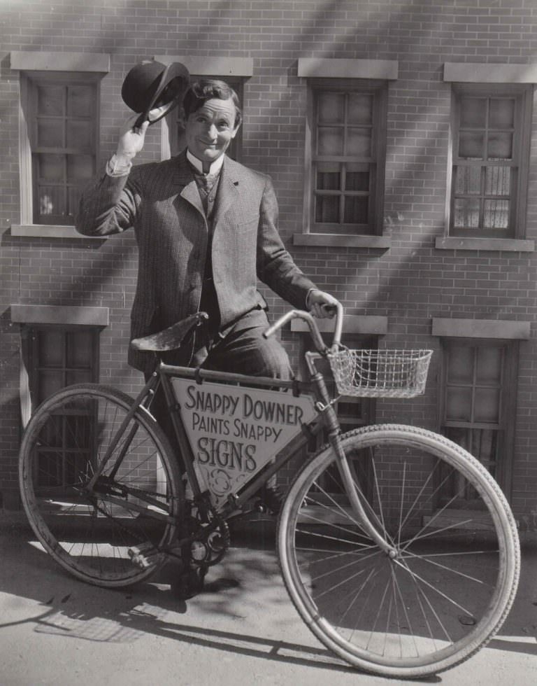 Roscoe Karns posing with a bike, tips his hat.