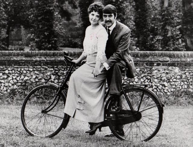 Pauline Collins and John Alderton posing on a bike.